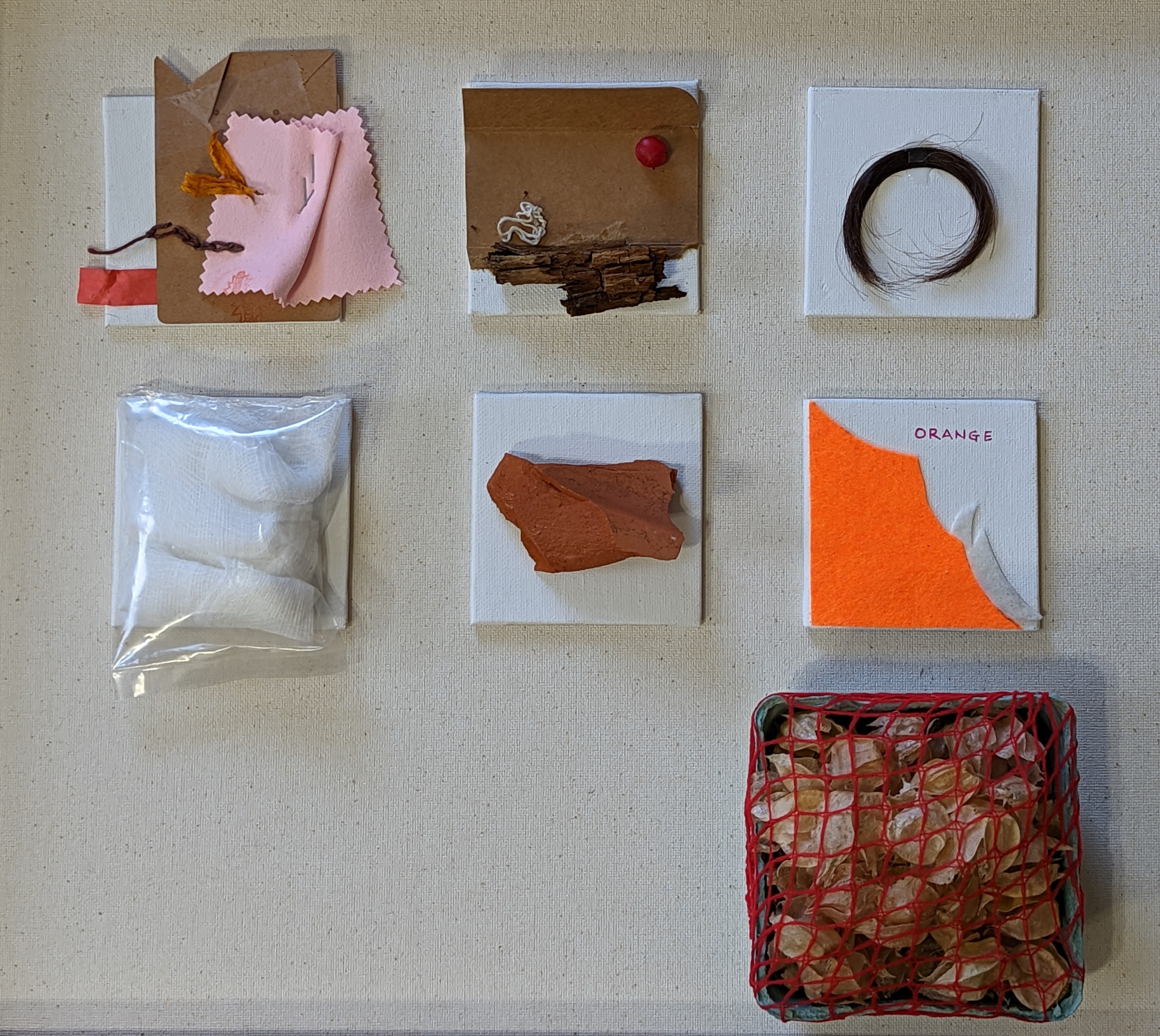 Grid of 6 small mixed-media assemblages on canvas on a linen wall: fabric swatches with orange yarn fringe, weathered wood bark with white thread loop, coiled dark hair ring on canvas, white garment in clear plastic bag, terracotta painted clay fragment, and bright orange felt triangle with 'ORANGE' text, with a red-net basket filled with dried seed pods below right.