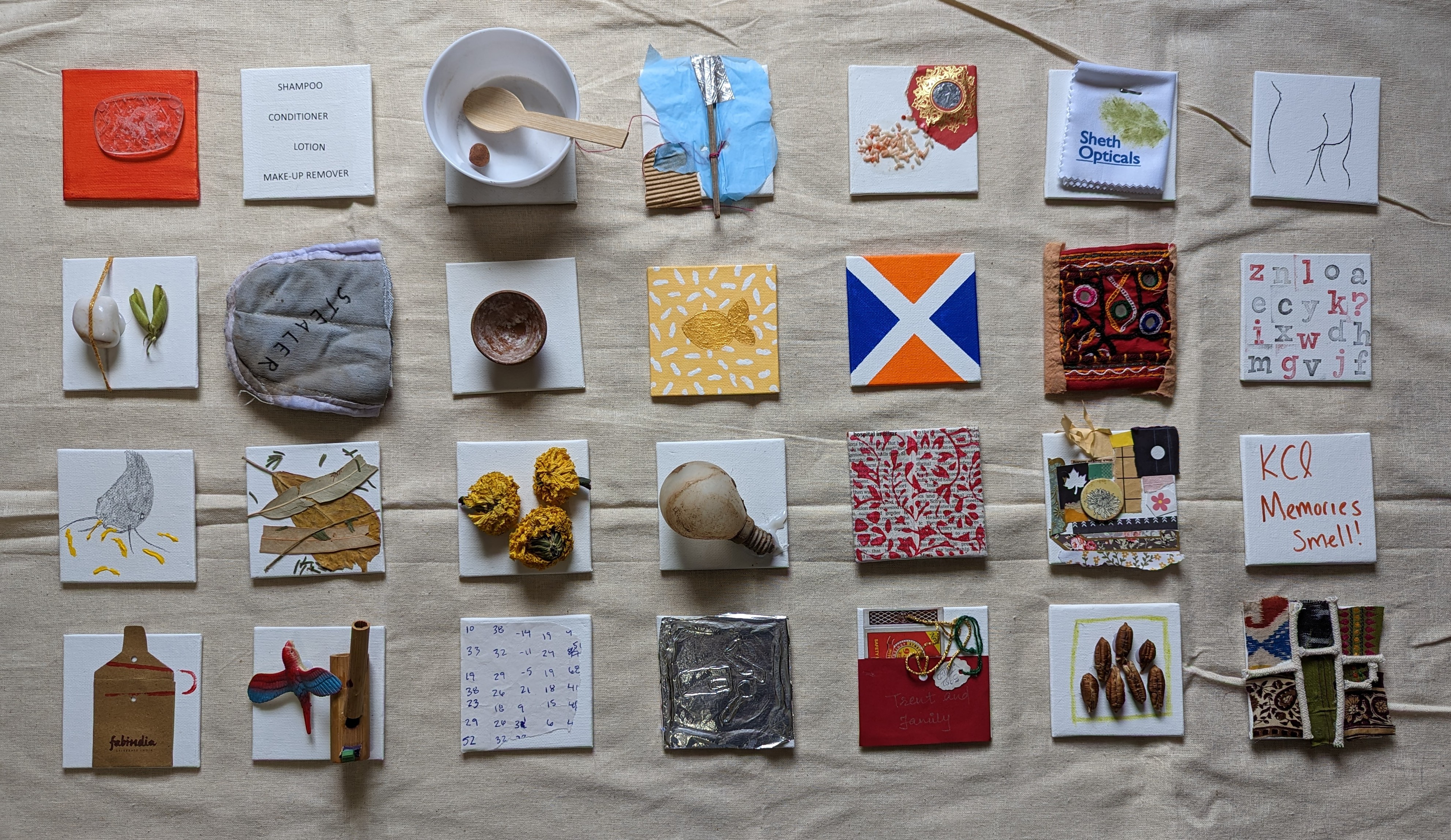 Dense wall installation of approximately 24 mixed-media assemblages on linen: painted panel with toiletry list 'SHAMPOO / CONDITIONER / LOTION...', white bowl with spoon and seed, tissue butterfly with scissors, 'Sheth Opticals' fabric scrap, body silhouette drawing, embroidered Gujarati textile square, and cut-letter alphabet grid across multiple rows.