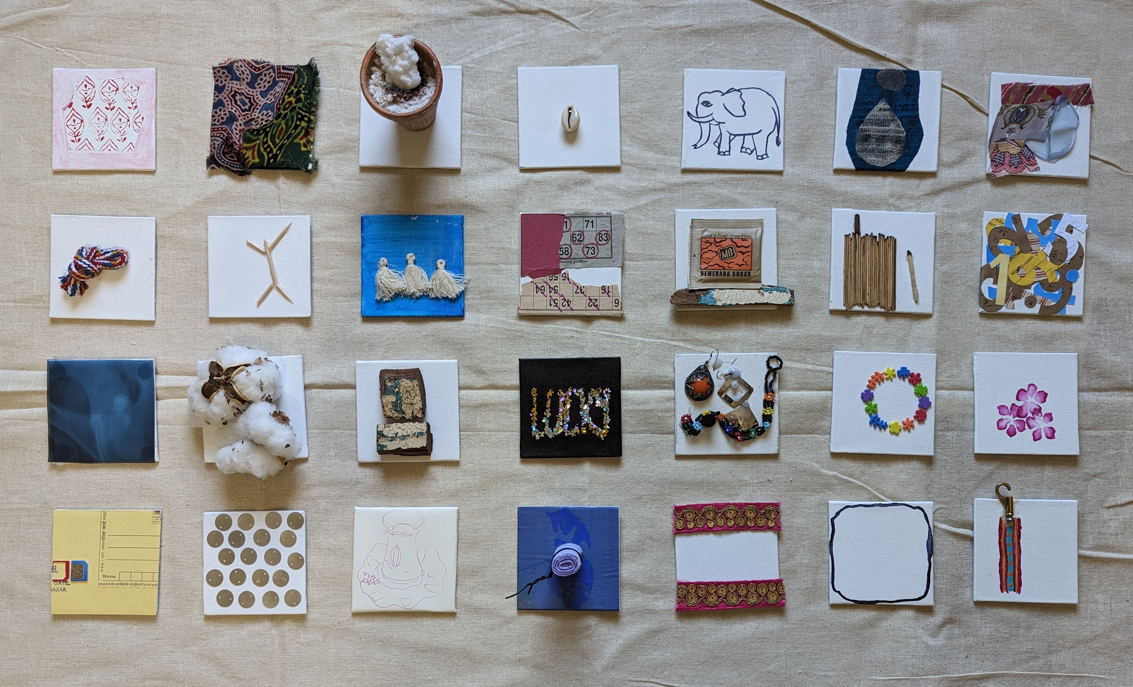 Wall installation of approximately 28 small assemblages in 4 rows on linen: floral block-printed fabric, terracotta pot with cotton bolls, cowrie shell on canvas, ink-drawn elephant, denim teardrop, twisted rope, crossed twigs, sugar sachet collage, bundle of matchsticks, sequined blue stitched box, flower wreath ring, rhinoceros contour drawing, and parasol charm.