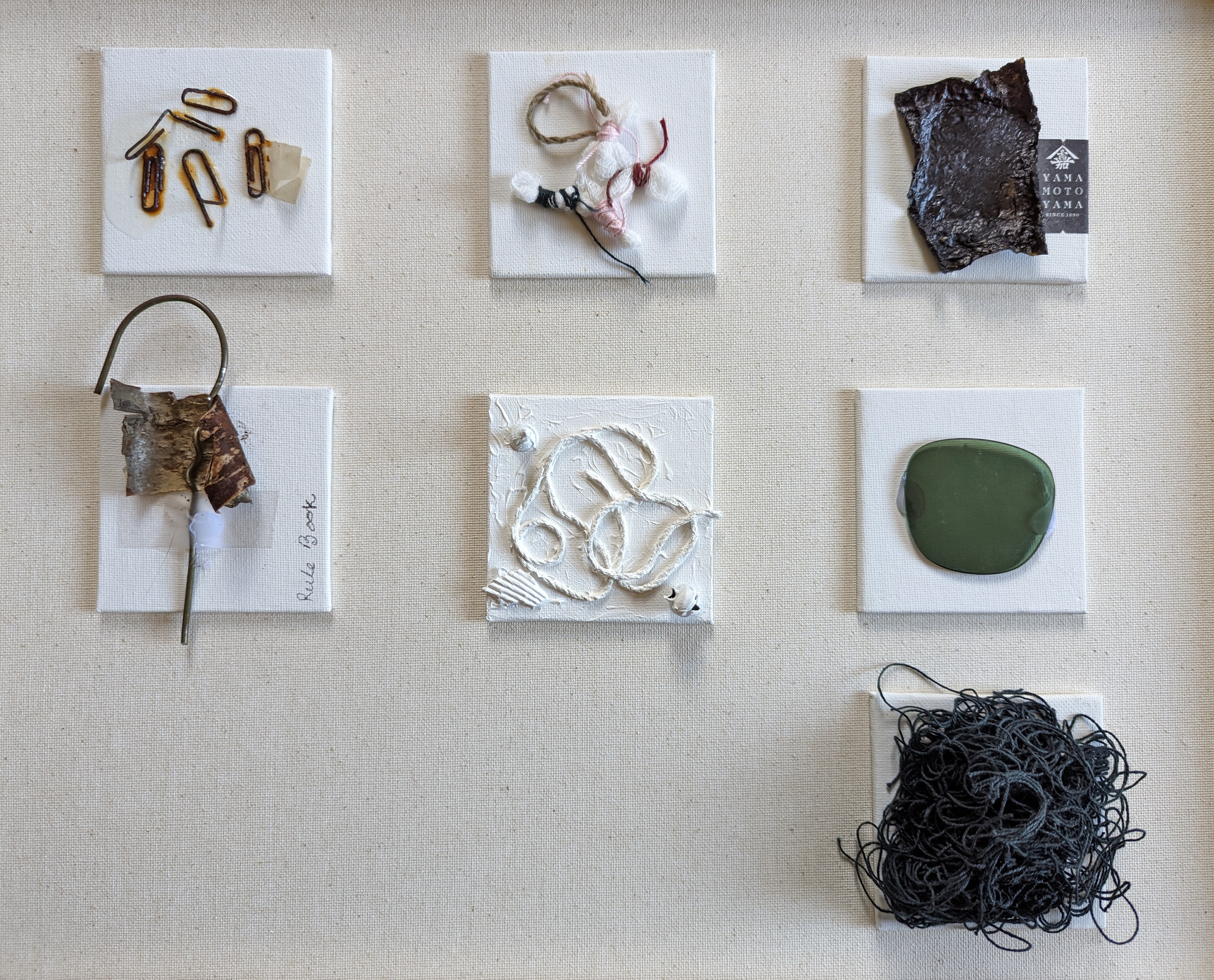 Grid of 7 small assemblages on linen: rusted safety pins on canvas, knot of multicolored threads, dark seaweed or bark with a 'YAMAMOTO YAMA' tea label, bent wire stem with birch piece labeled 'Rule Book', white gesso relief with looping cord, smooth green oval stone, and a tangled ball of dark charcoal-grey thread.