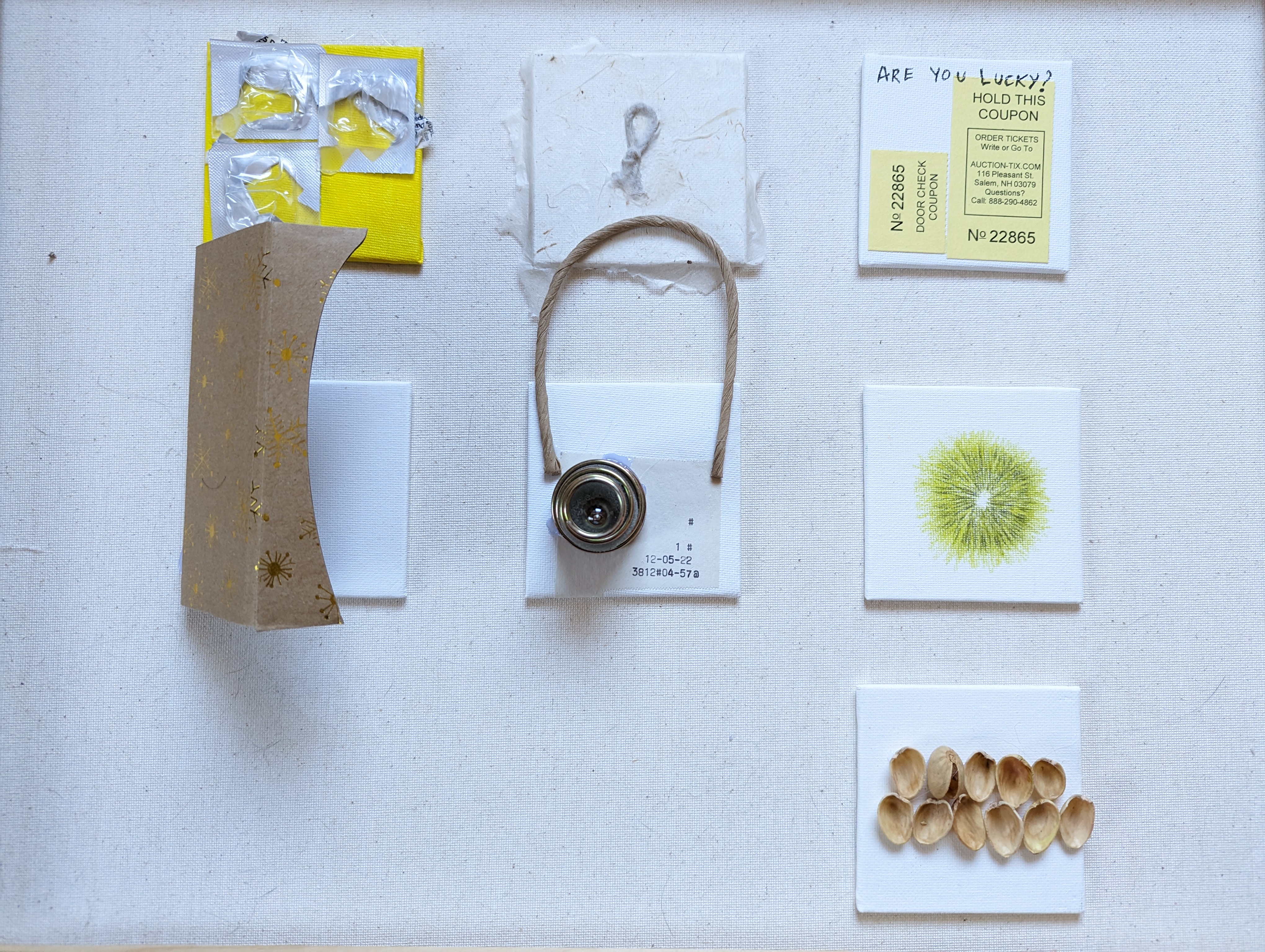 Grid of small assemblages on white linen: yellow and gold package with pill blister packs, thick textured handmade paper with twine loop and metal peephole lens stamped with date code, yellow door coupon 'ARE YOU LUCKY? HOLD THIS COUPON No 22865', fine pen dandelion illustration, and an arched row of hollow pistachio shell halves.